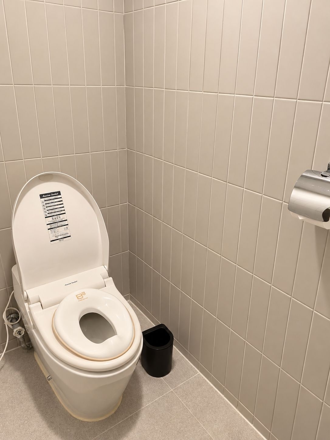 Une salle de bain standard équipée de toilettes, d’une poubelle et d’un porte-rouleau de papier toilette fixé au mur.