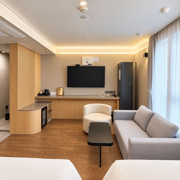 Una habitación de hotel moderna con sofá, una mesita, televisor montado en la pared, refrigerador y un separador de madera.