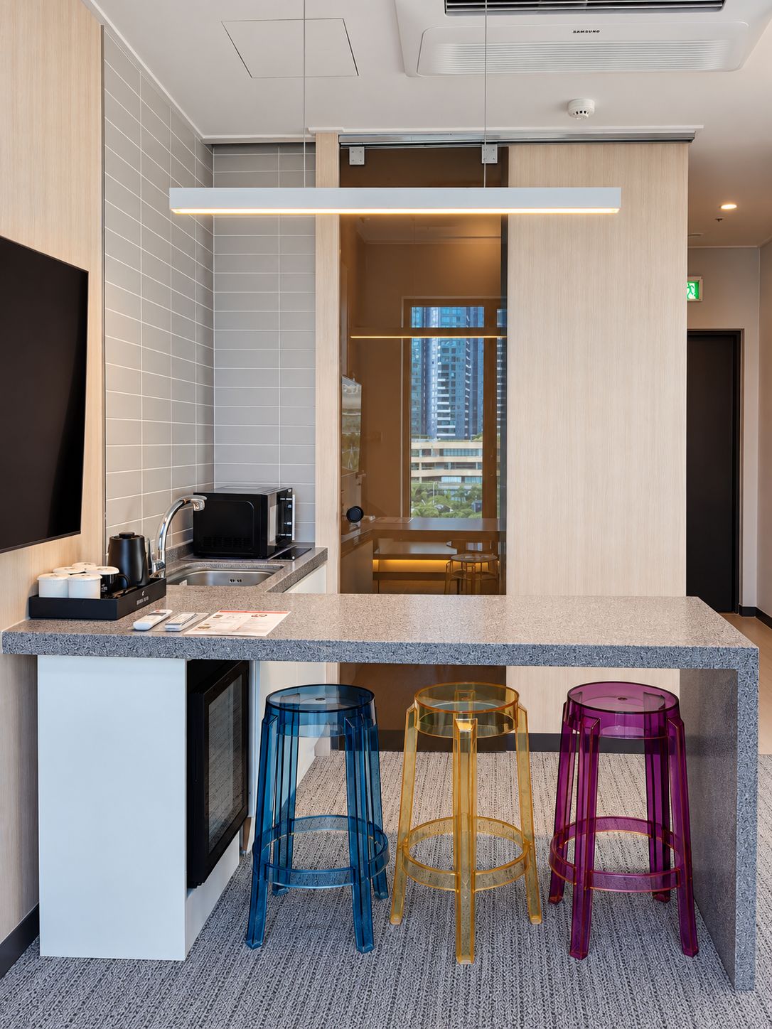 A modern kitchen with a built-in microwave, a coffee machine, and a sink. There are colorful stools and a window with a view of buildings.