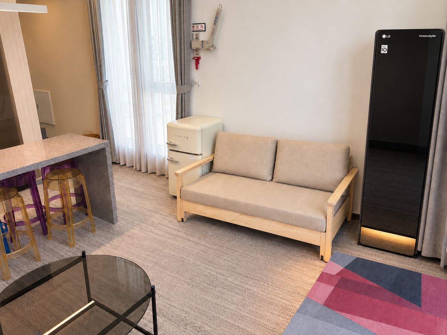 A modern living room with a sofa, a small table, and a glass-top coffee table. There is a window with curtains, a radiator, and a black appliance on the wall.