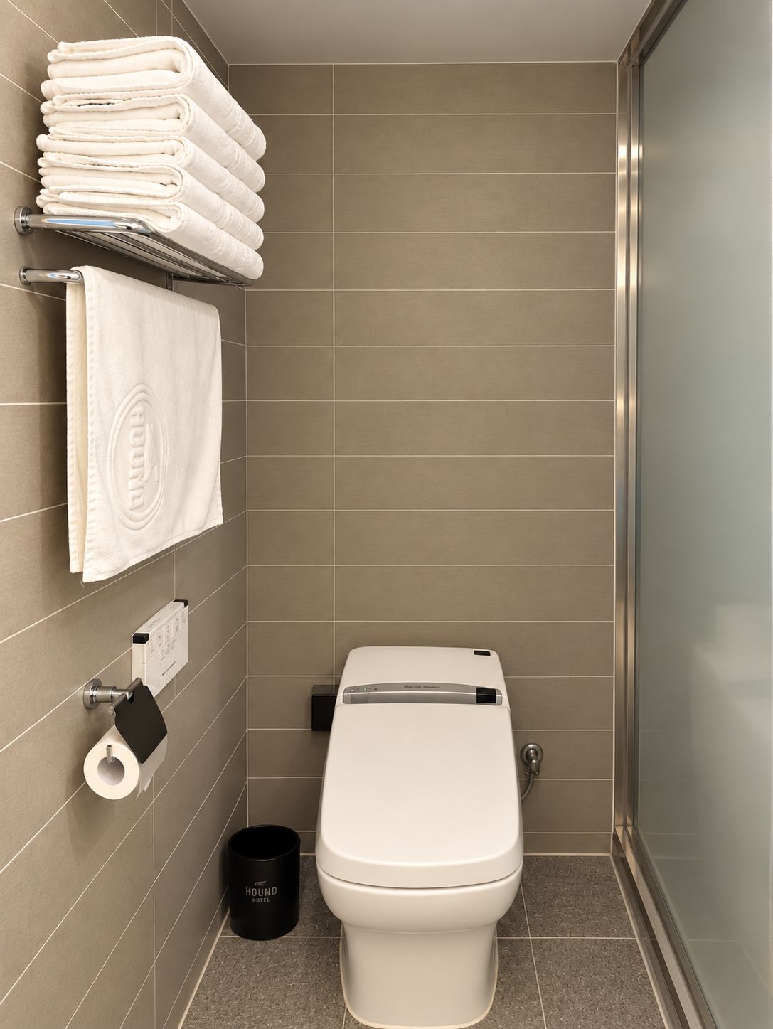A modern bathroom with a toilet, a towel rack with folded towels, a toilet paper holder, and a trash bin.