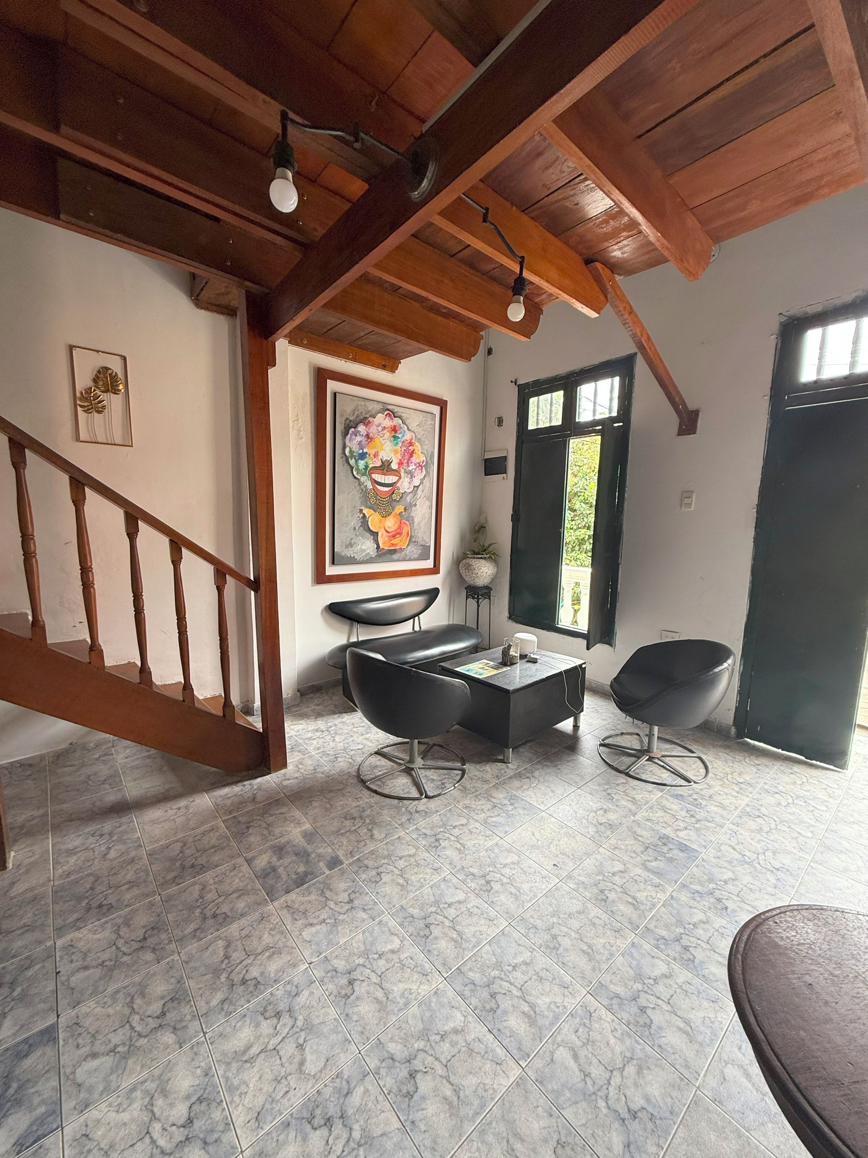 Una habitación con una escalera de madera, un sillón de cuero negro, suelo de baldosas de mármol, un cuadro en la pared y una ventana con vistas.