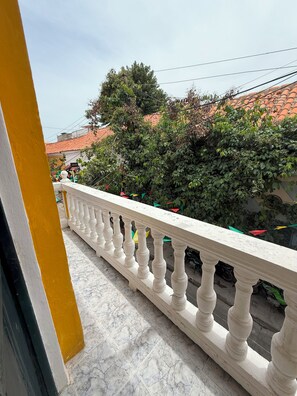 Un balcón con barandilla blanca, piso de baldosas y vista a un edificio con tejido rojo y vegetación.