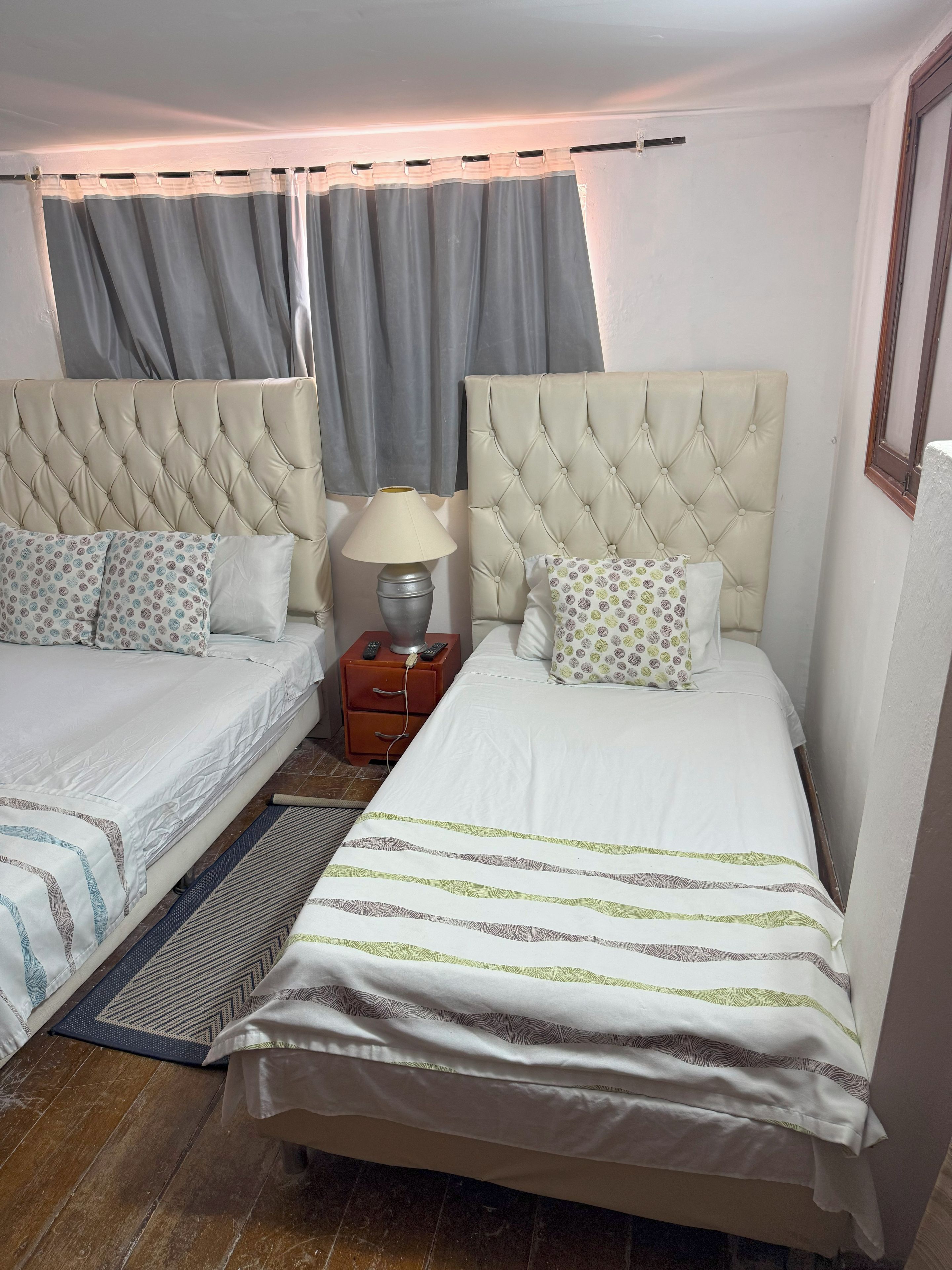 A hotel room with two beds, a nightstand, a lamp, and a window with curtains.