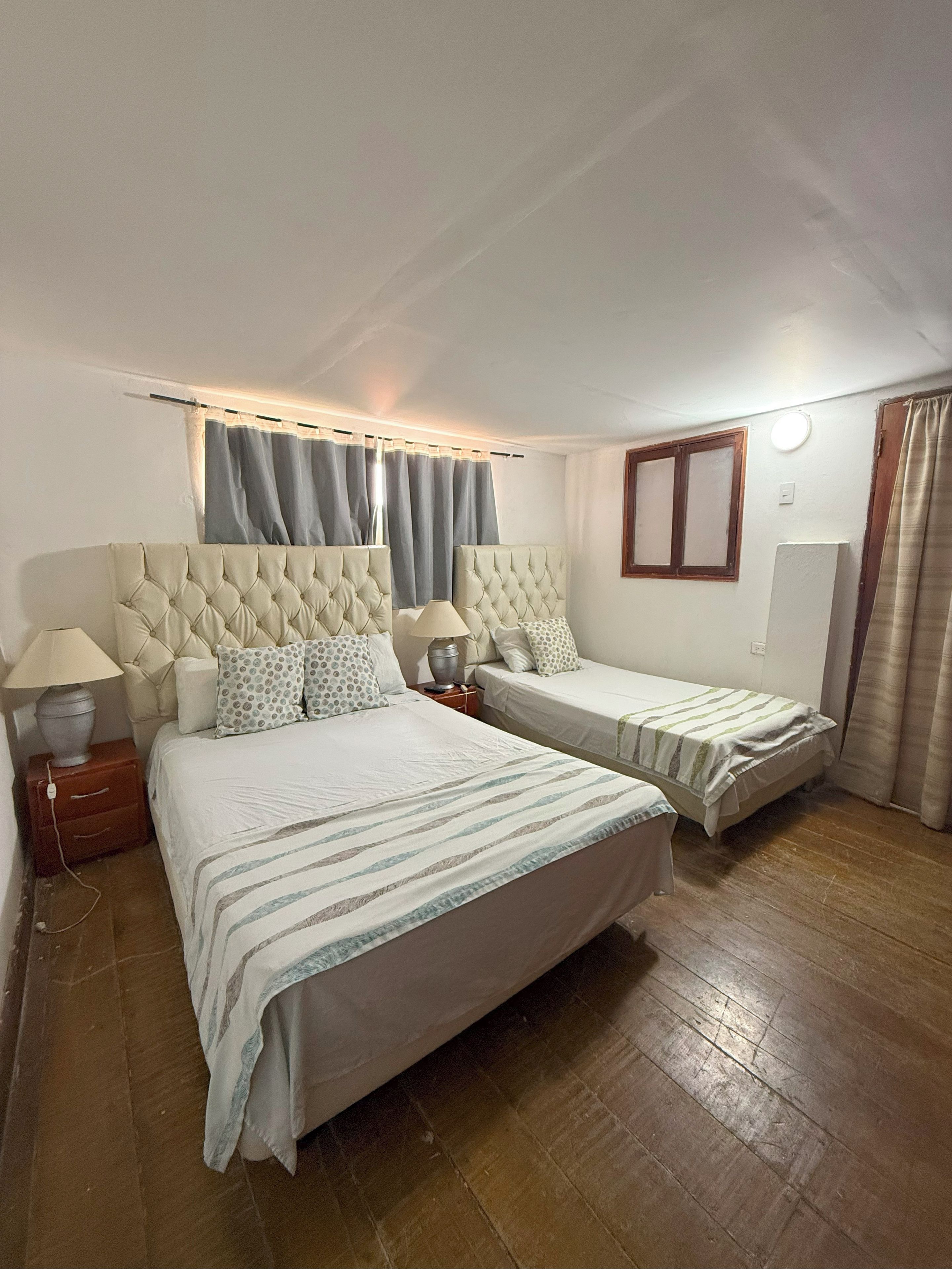 A bedroom with two beds, a window with curtains, a bedside table with a lamp, and a wall-mounted light.