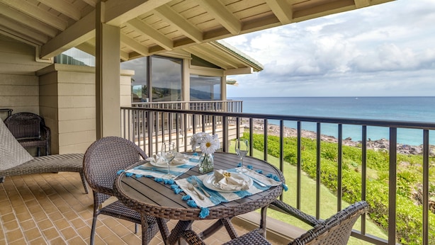 A covered outdoor dining area with wicker furniture, a table set for two, and a view of the ocean.