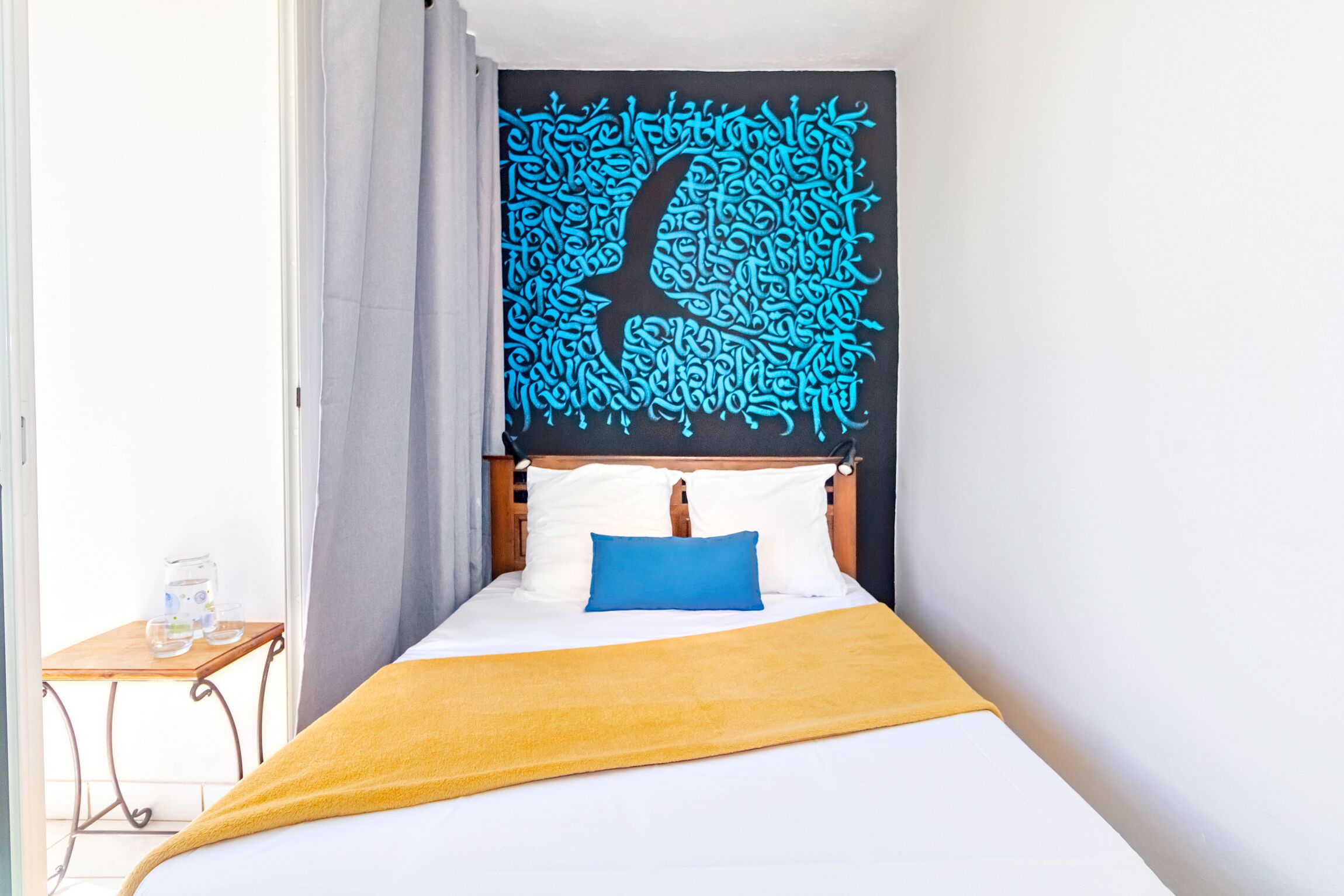 Une chambre à coucher avec un lit, une tête de lit en bois, un oreiller bleu, une couverture jaune et une tapisserie murale.