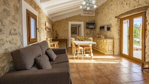 A cozy living room with a sofa, a dining table, and a kitchen area with stone walls and wooden cabinets. - Beautiful farm with 2 bedrooms, 2 bathrooms, wifi, pool and barbecue (Caimari)