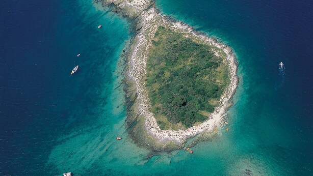Vista aérea de una pequeña isla con aguas turquesas cristalinas y algunas embarcaciones.