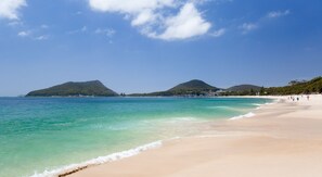 A beach with clear turquoise water, sandy shore, and hills in the background. - The Helm 5,22 Voyager Cl - waterfront unit (Nelson Bay)
