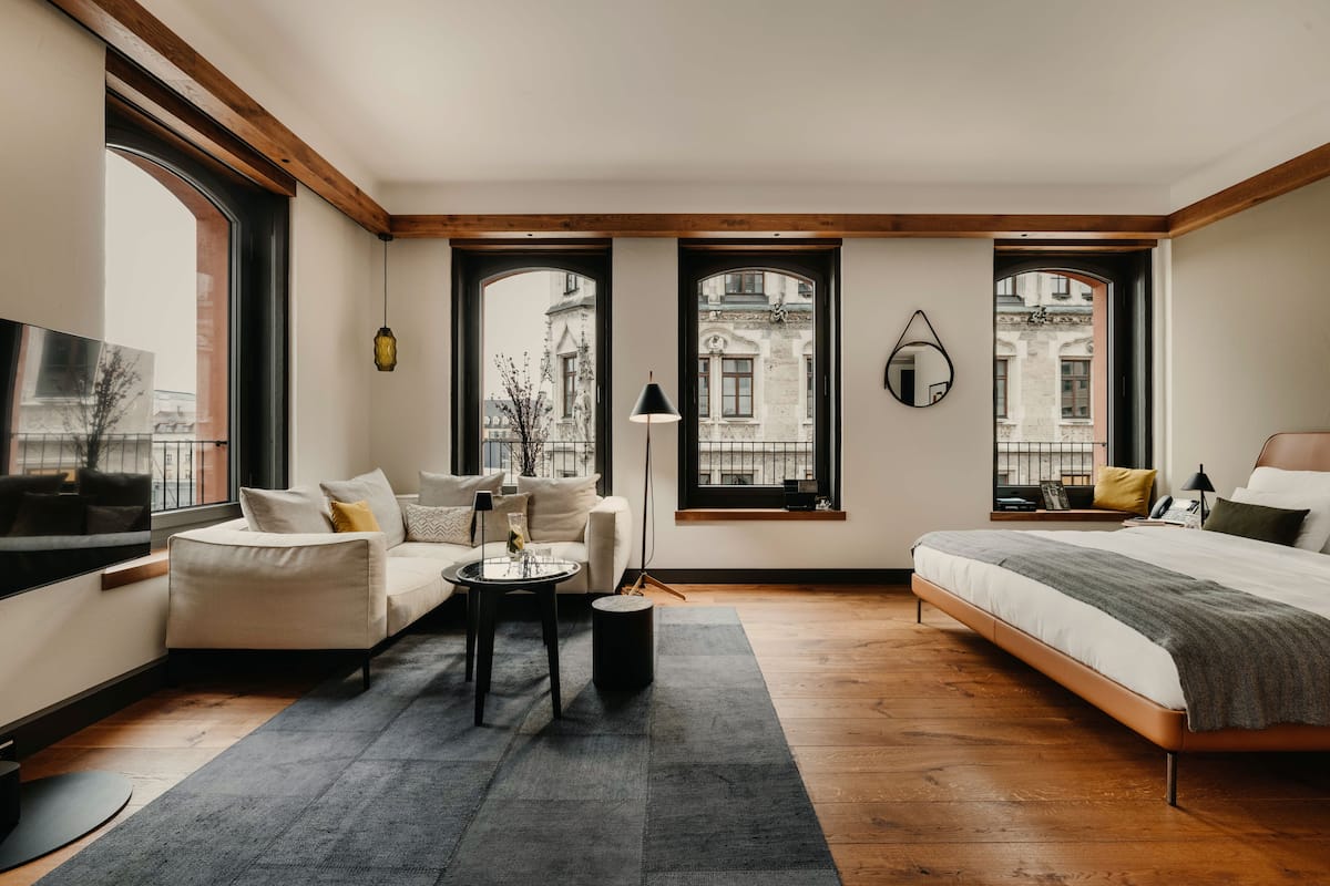 A modern hotel room with a sofa, a small table, a bed, and large windows offering a view of the city.
