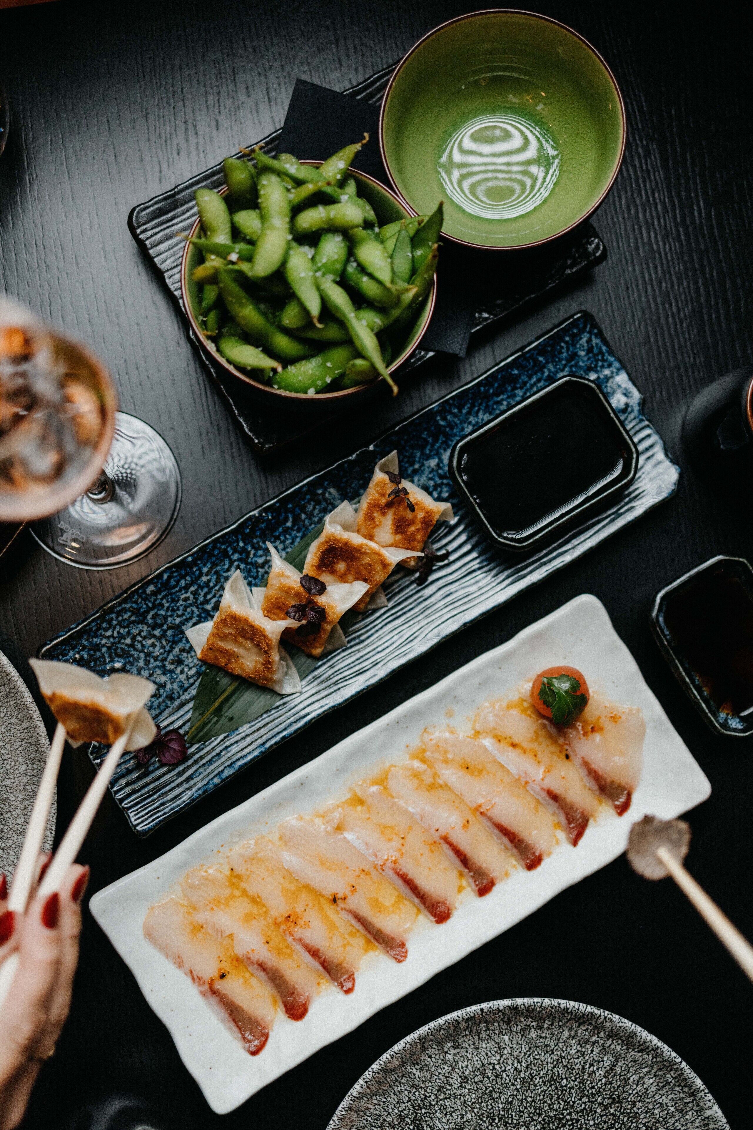 Ein Esstisch mit verschiedenen Gerichten, darunter Sushi, Edamame und Spieße, sowie kleinen Saucenschalen.