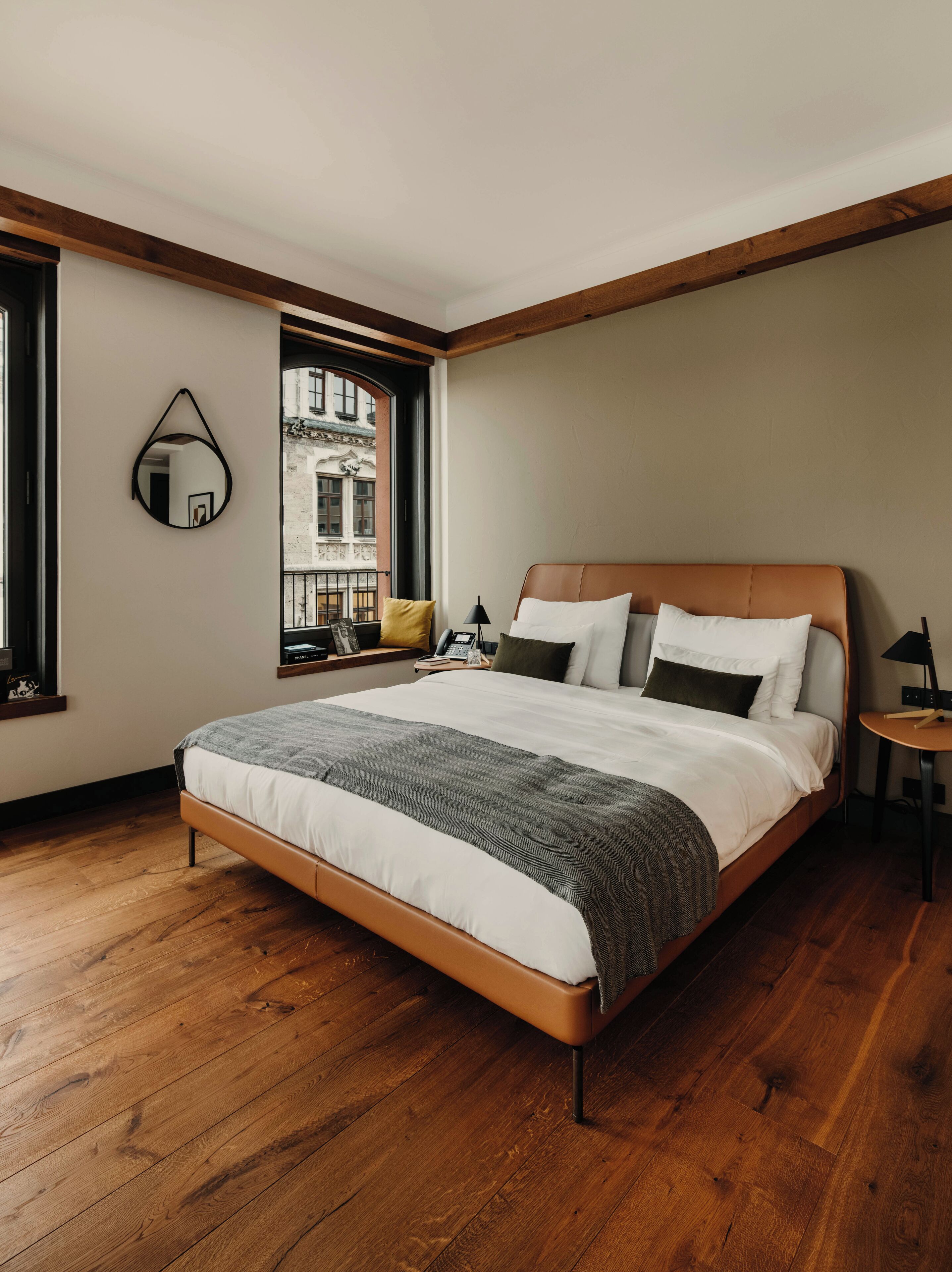 Un dormitorio moderno con piso de madera, una cama grande con cabecera, una mesita de noche, un espejo y una ventana con vista a edificios.