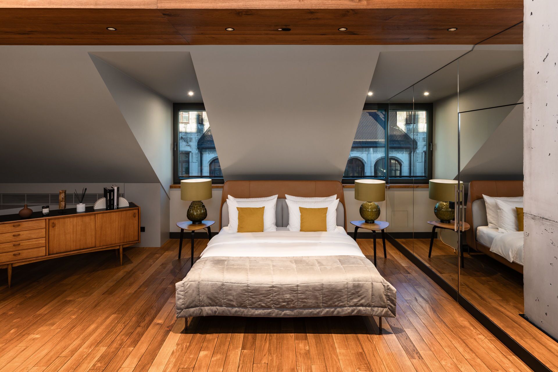 Una camera da letto moderna con un letto grande, due comodini con lampade, una cassettiera in legno e vista su un edificio attraverso una finestra.