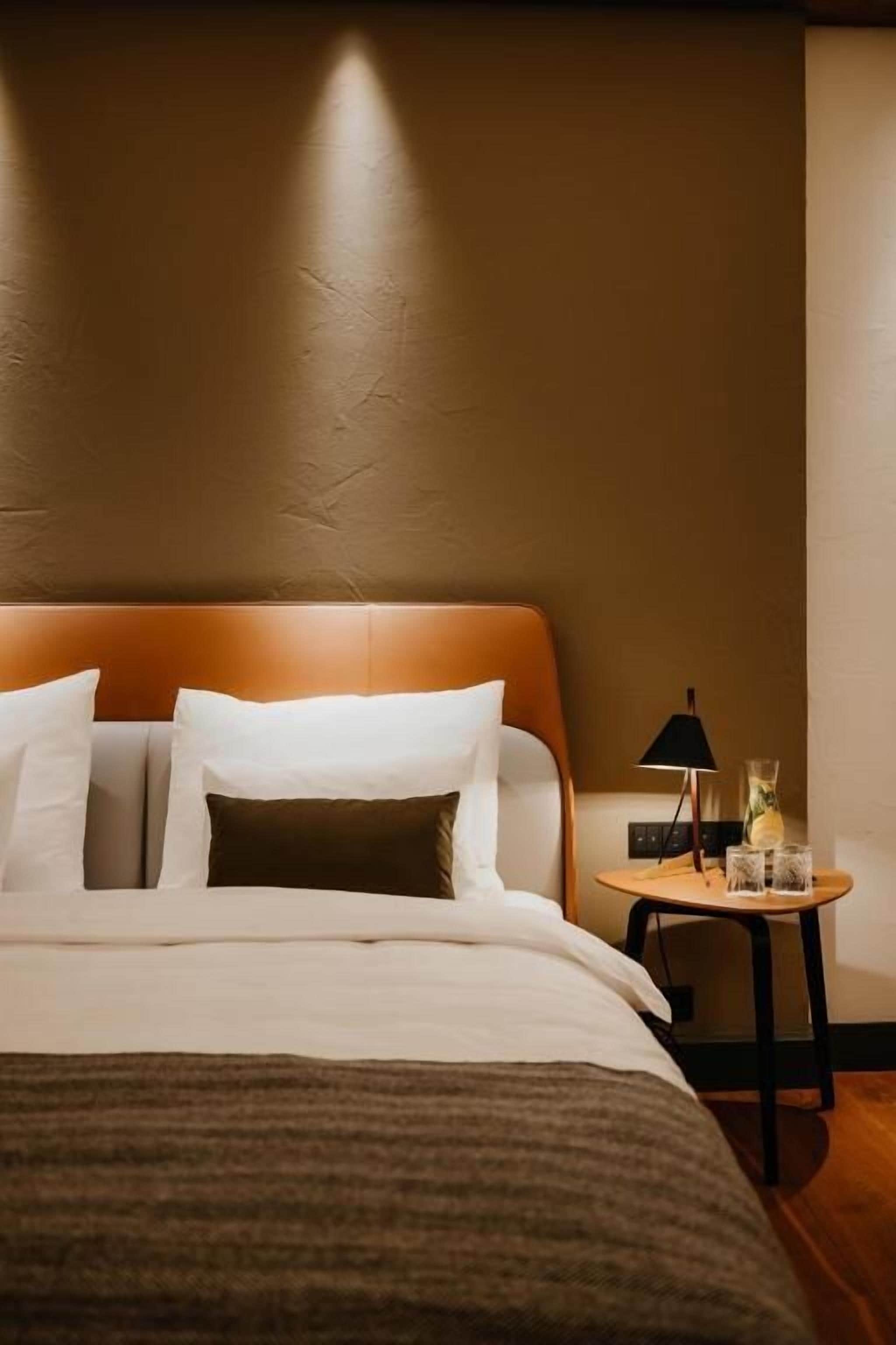 A neatly made bed with white and brown bedding, a wooden headboard, and a small bedside table with a lamp and glasses.