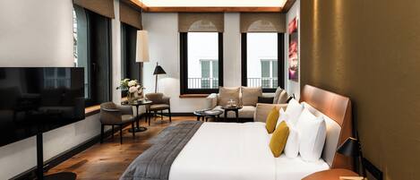 A modern hotel room with a large bed, a TV, a sofa, a small table, and a balcony with a view.