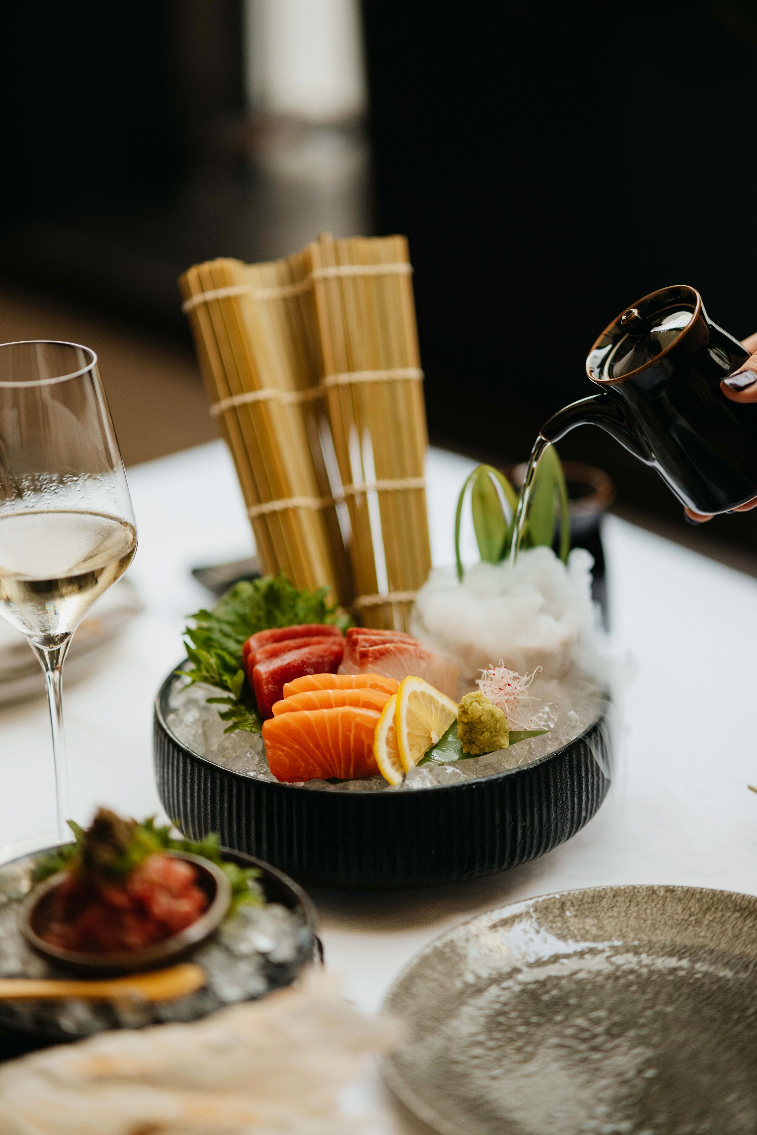 A sushi platter with various types of fish, garnishes, and a glass of white wine on a table.