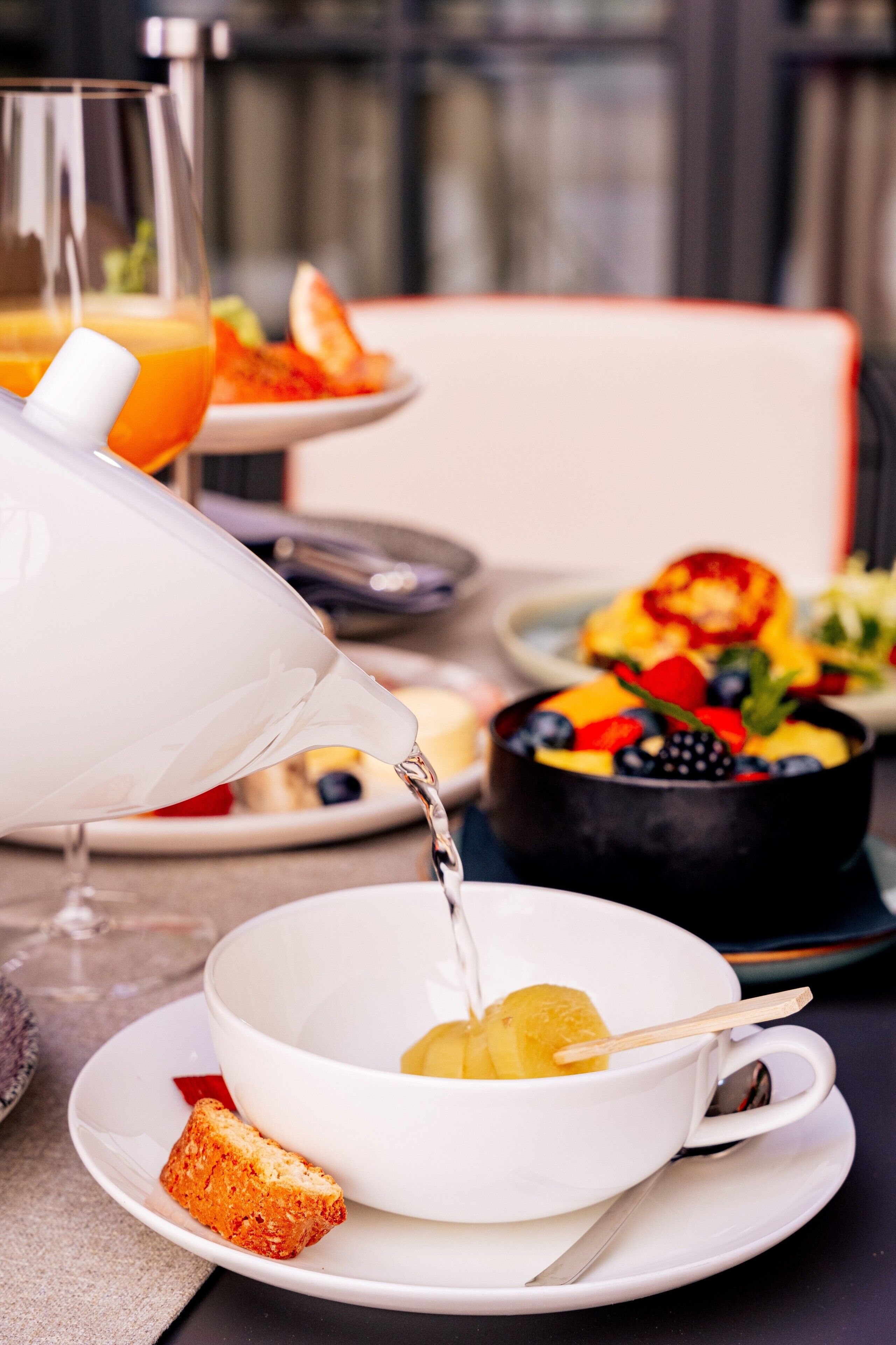 Una escena de desayuno con una jarras vertiendo, un bol de fruta y un vaso de jugo de naranja sobre una mesa.