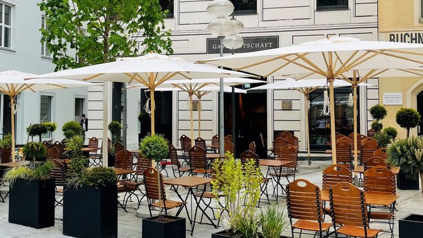 An outdoor cafe area with tables and chairs, surrounded by potted plants and umbrellas. - DO & CO Hotel München (Munich)