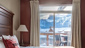 Ein Hotelzimmer mit Bett, Nachttisch, Lampe und Blick auf verschneite Berge durch große Fenster.