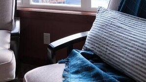 Ein Zimmer mit großem Fenster, einem Bett mit blauer Decke und einem Sessel.
