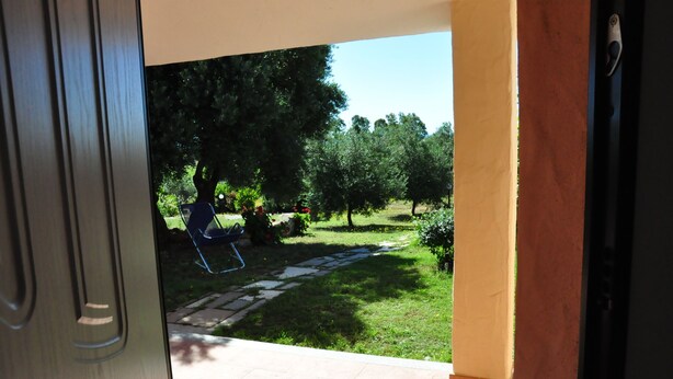 An open door leading to a patio with a lawn, a stone pathway, and a blue chair. - Sa Teria Village - apartment 2 (tertenia)