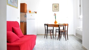Een ruimtelijk ingerichte leefruimte met een rode bank, een eettafel met stoelen, een keukenhoek met een koelkast en een klein raam.