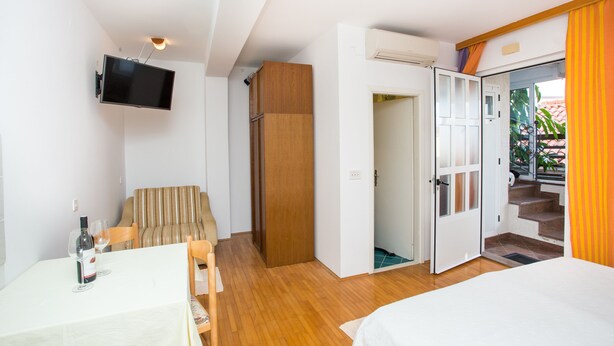 Une chambre à coucher avec un lit, un téléviseur fixé au mur, une table à manger avec des chaises et une porte en bois donnant sur un escalier.