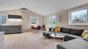 A modern kitchen with a built-in oven, a dining area with a table and chairs, a grey sofa with yellow cushions, and large windows offering a view of the outdoors.
