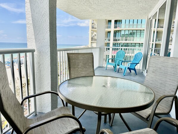 A balcony with a glass table and chairs, overlooking a beach and a building with multiple balconies.