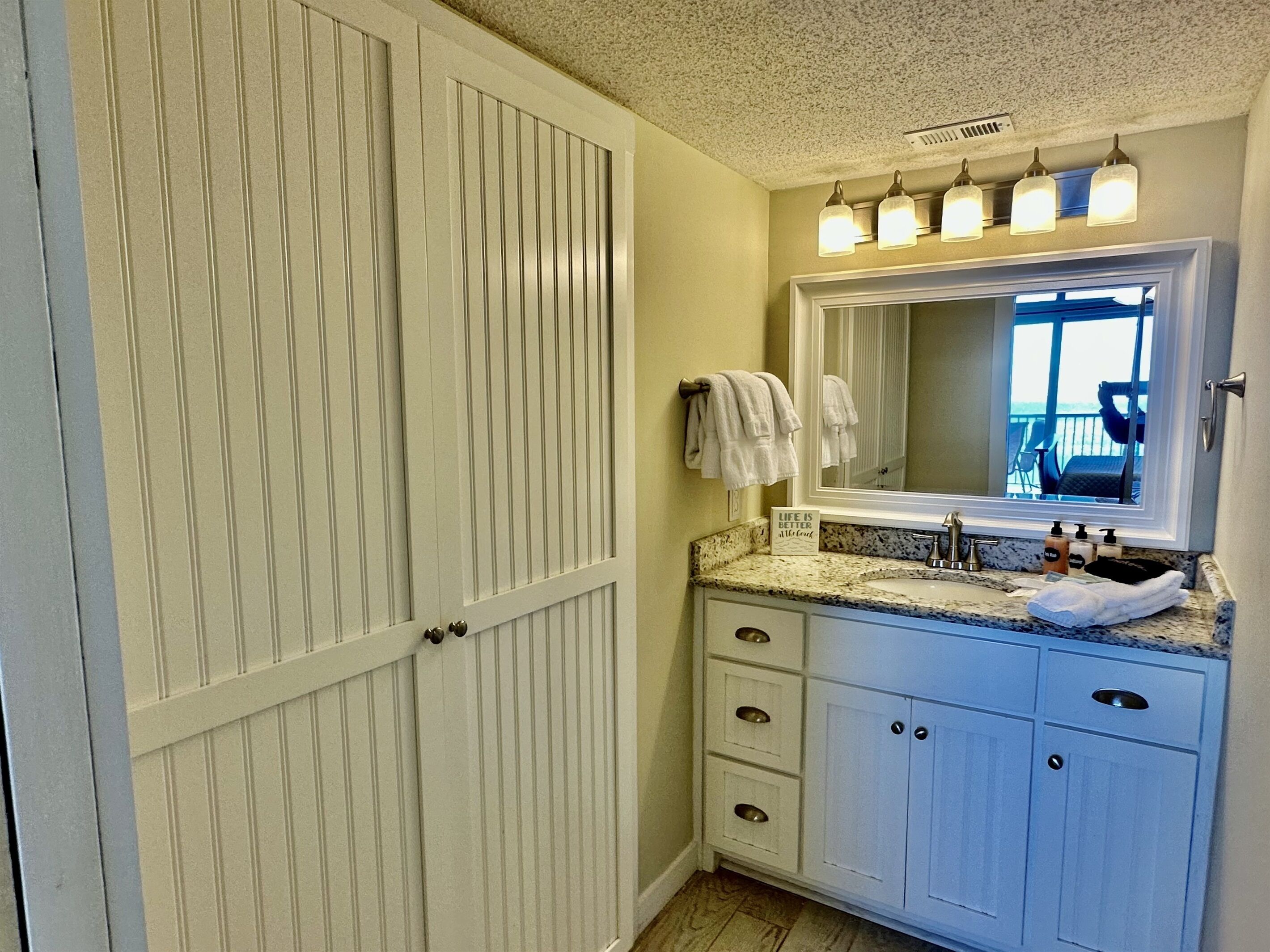 A bathroom with a large mirror, a sink, and a vanity with drawers. There are towels hanging on the wall and a sign with a quote.