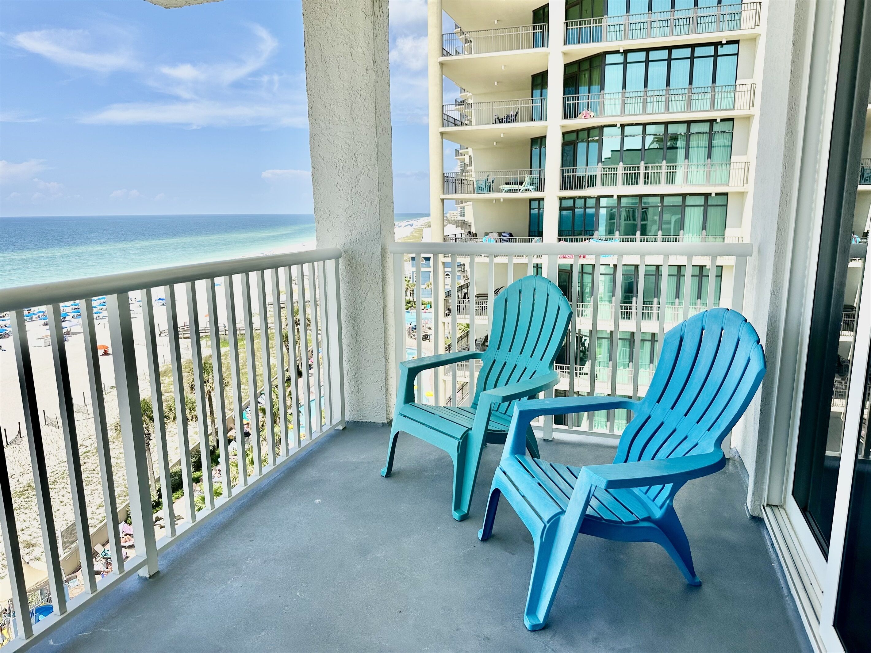 A balcony with two blue chairs overlooking a beach and a building with multiple balconies.