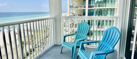 A balcony with two blue chairs overlooking a beach and a building with multiple balconies.