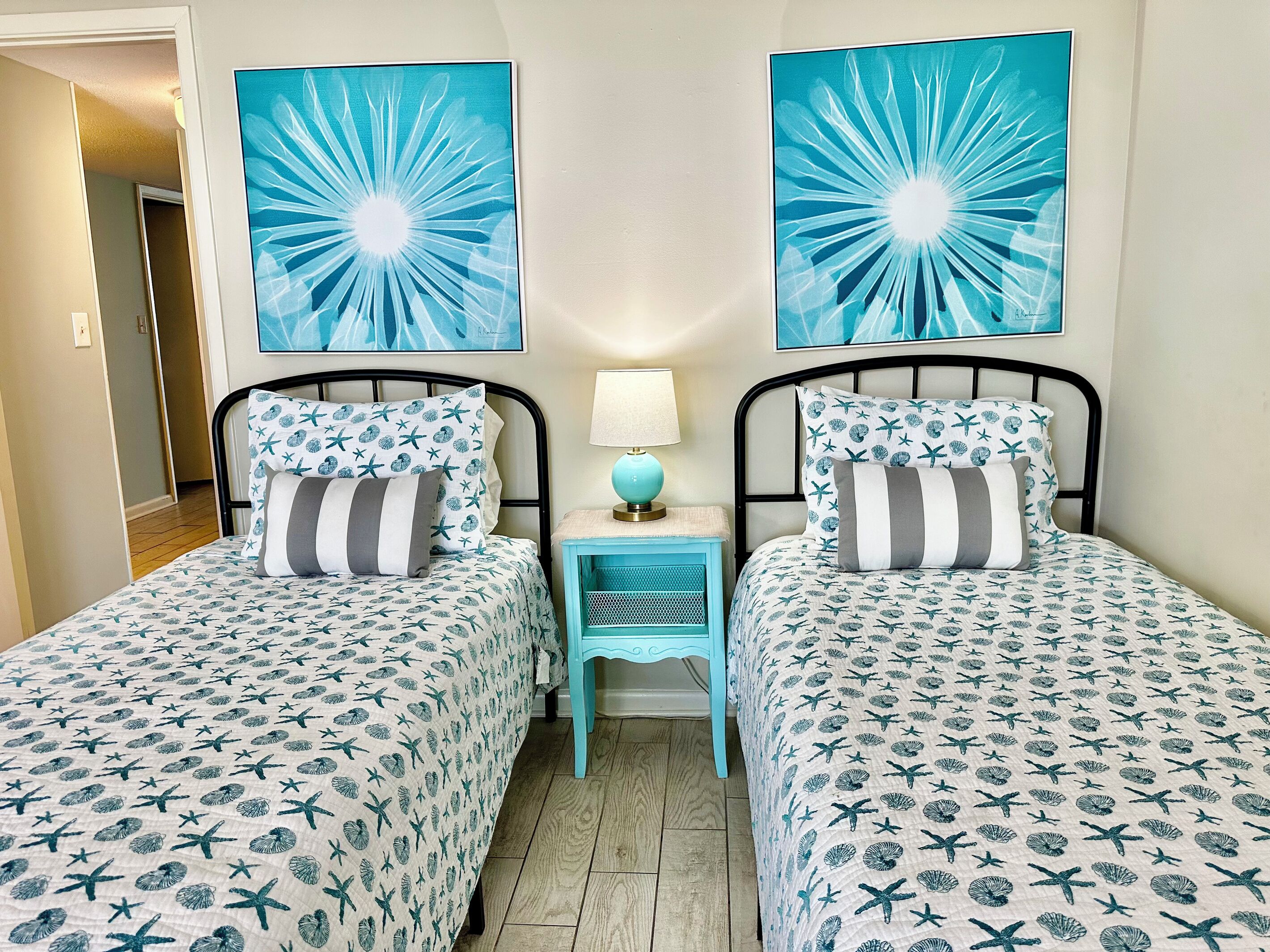 Una habitación con dos camas, cada una con una colcha y almohadas estampadas, una mesita de noche con lámpara, y dos grandes cuadros abstractos en la pared.