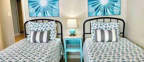 Una habitación con dos camas, cada una con una colcha y almohadas estampadas, una mesita de noche con lámpara, y dos grandes cuadros abstractos en la pared.