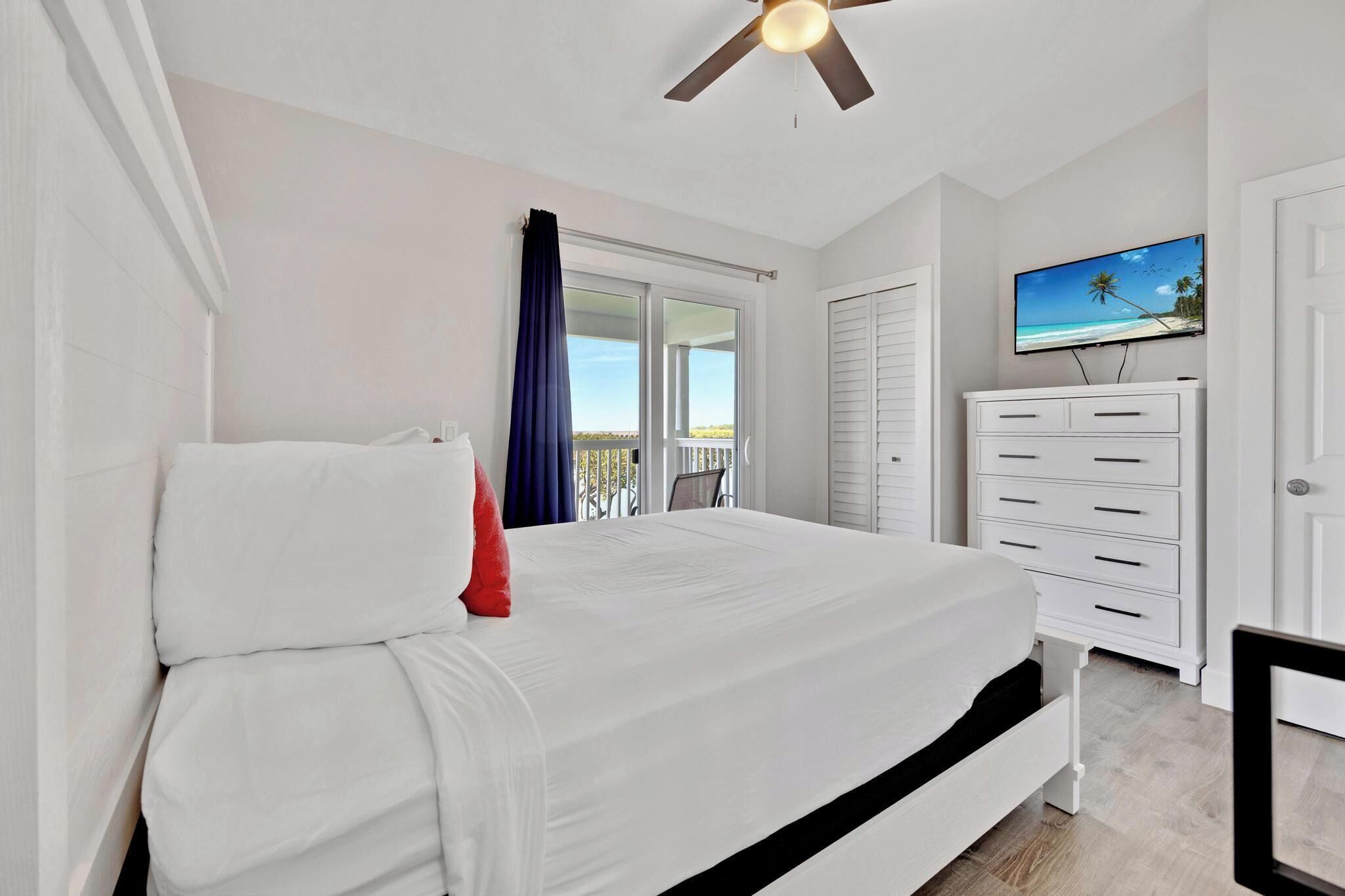 A bedroom with a bed, a dresser, a television, and a balcony with a view of the beach.