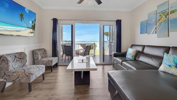 Een woonkamer met een bank, twee fauteuils, een salontafel en een plafondventilator. Er is een groot raam met uitzicht op het strand en een terras met tuinmeubilair.