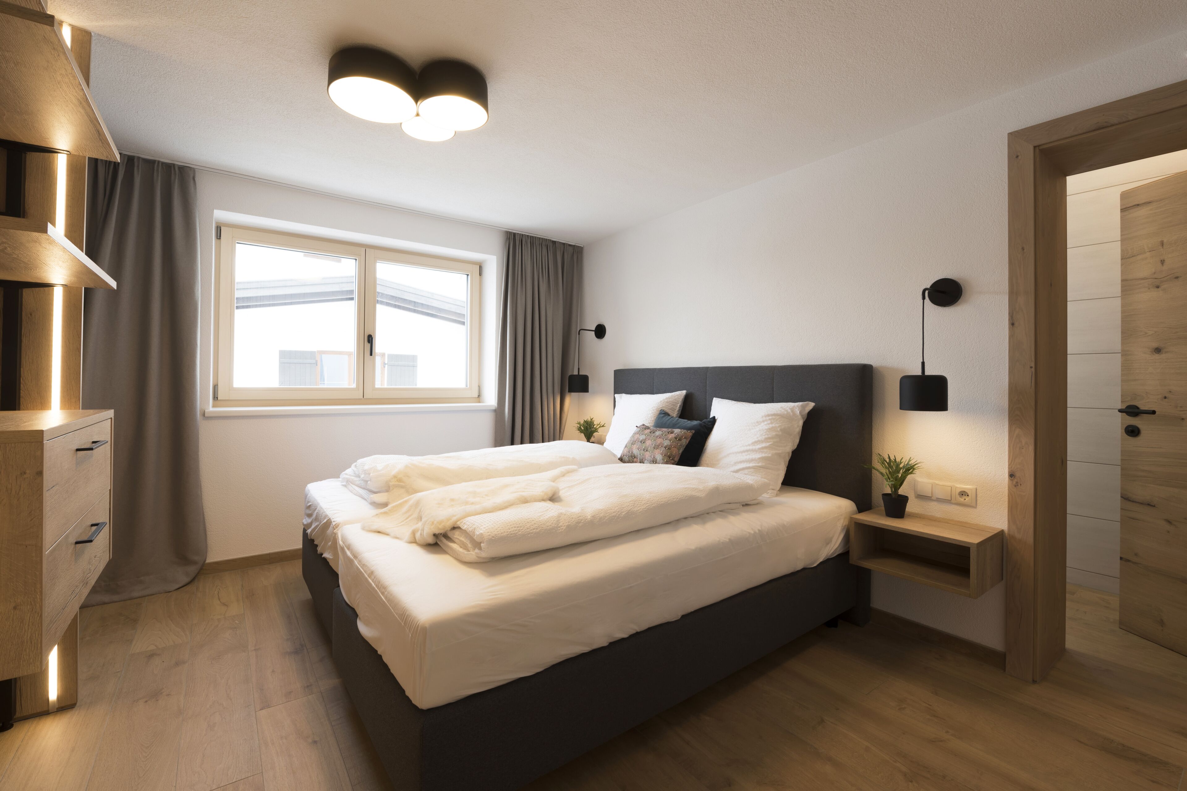 Ein modernes Schlafzimmer mit einem großen Bett, Nachttischen, einem Fenster mit Vorhängen und einer Holztür.
