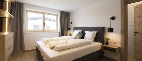 A modern bedroom with a large bed, bedside tables, a window with curtains, and a wooden door.