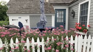 A white picket fence with pink flowers in front of a house with a gray roof and a blue door. - OCEAN VIEWS & A/C in cozy condo w/upscale finishes!- STEPS TO PRIVATE BEACH (Mashpee)