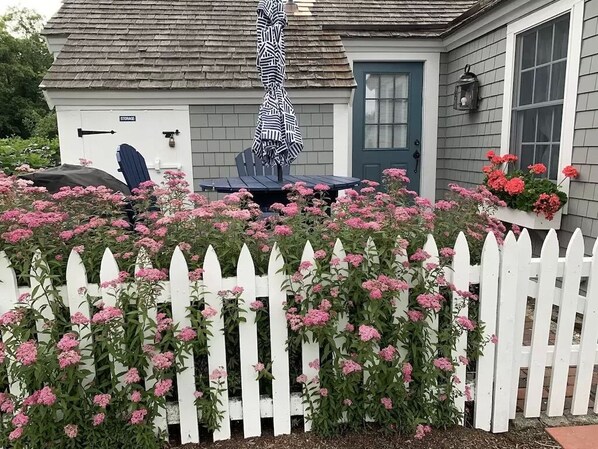 A white picket fence with pink flowers in front of a house with a gray roof and a blue door. - OCEAN VIEWS & A/C in cozy condo w/upscale finishes!- STEPS TO PRIVATE BEACH (Mashpee)