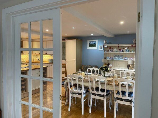 A dining area with a wooden table, white chairs, and a kitchen in the background. - Attractive former stables located on beautiful Victorian private country estate (Rousdon)