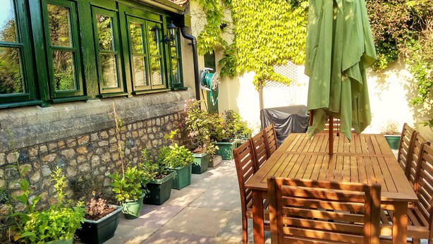 Une table et des chaises en bois sont disposées à l’extérieur, accompagnées de plantes en pot et d’un parasol vert.
