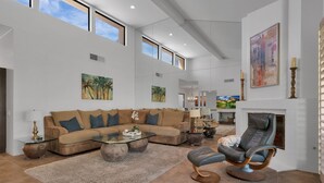 A spacious living room with a large sectional sofa, a glass coffee table, and a fireplace. There are multiple windows providing natural light, and artwork is displayed on the walls. - PGA Condo with Gorgeous View of Mountains and Fairway (La Quinta)