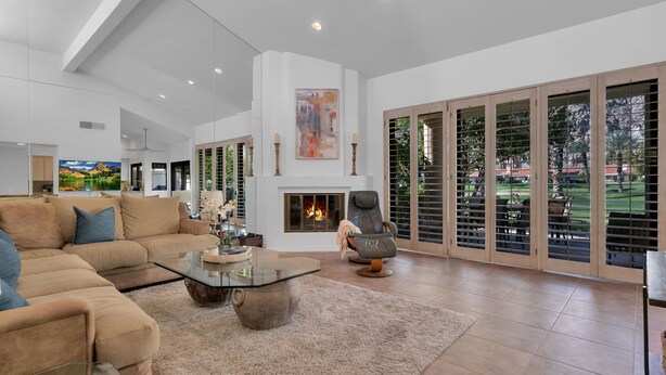 A spacious living room with a large sofa, a glass coffee table, a fireplace, and a view through large sliding doors to an outdoor area. - PGA Condo with Gorgeous View of Mountains and Fairway (La Quinta)