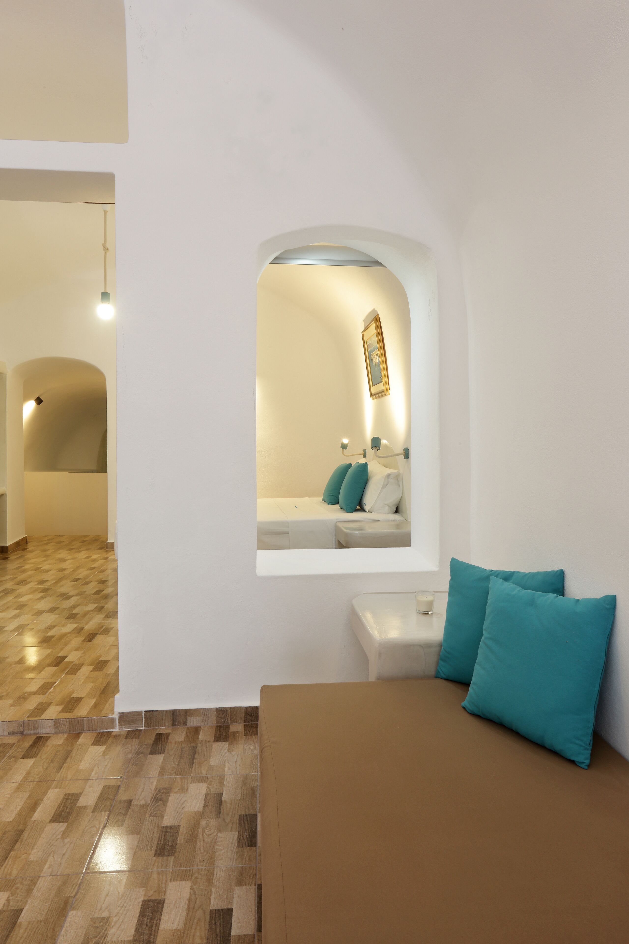 A hotel room with a brown sofa, teal pillows, a small window with a view of a bathroom, and a checkered floor.