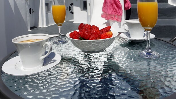 Ein Tisch mit Frühstücksangeboten, darunter eine Schale Erdbeeren, ein Glas Orangensaft, eine Tasse Kaffee und ein weiteres Glas Orangensaft. Der Tisch steht draußen mit Blick auf weiße Gebäude und einen klaren, blauen Himmel.