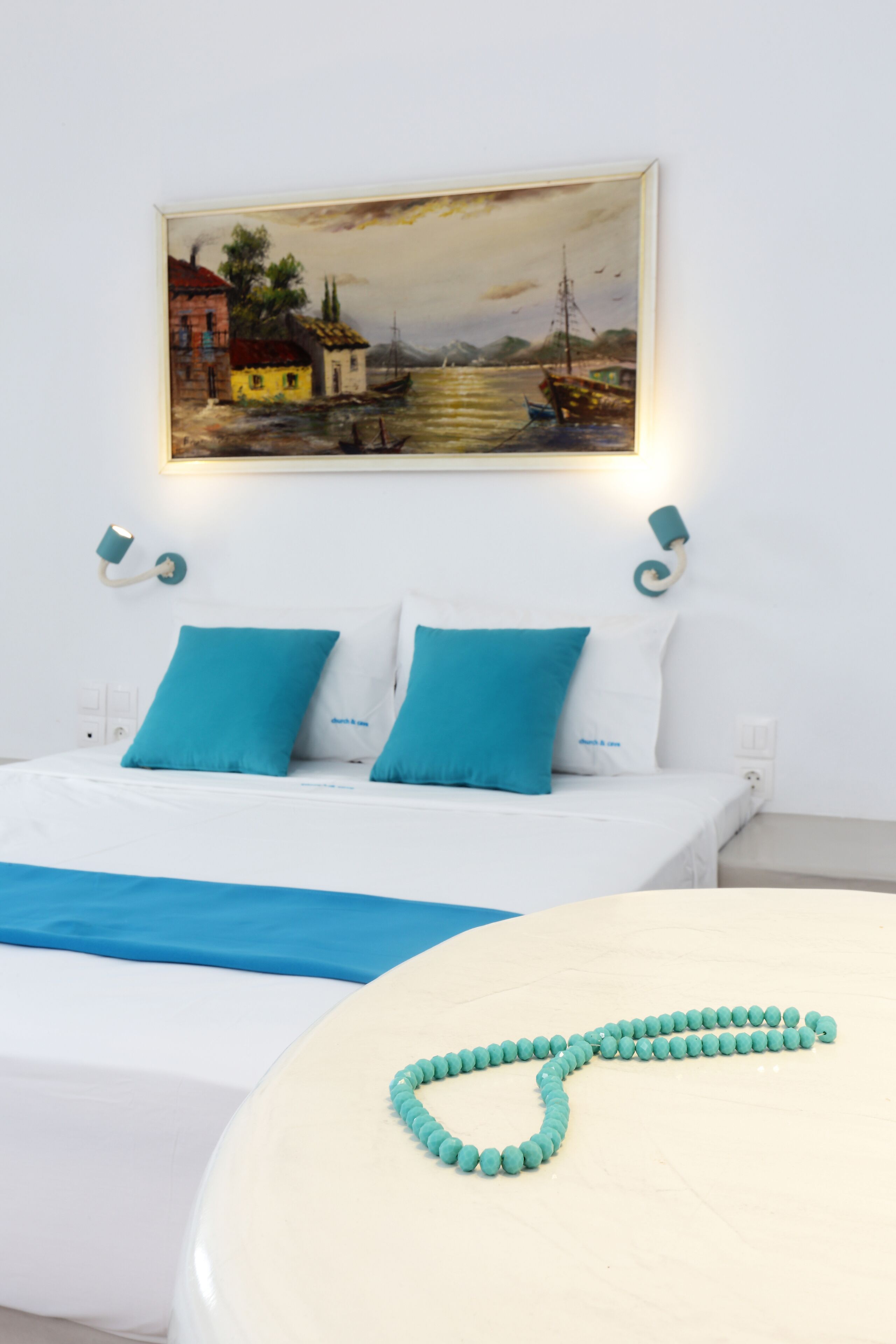 A hotel room with a bed, a painting of a coastal scene, and a teal beaded necklace on the bedside table.