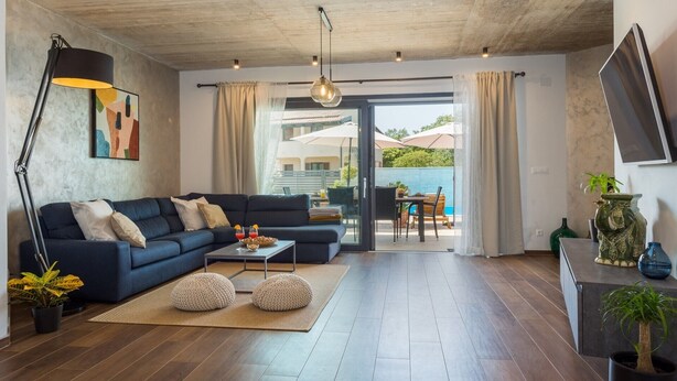 A modern living room with a dark blue sofa, a coffee table, and a floor lamp. There is a sliding glass door leading to an outdoor patio area with a table and chairs. - Modern Villa Leonardo with an outdoor pool (Porec)