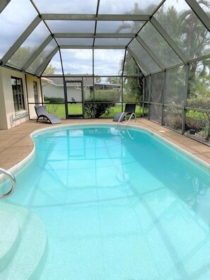 A covered swimming pool with two lounge chairs, a glass door leading outside, and a view of the surrounding greenery.